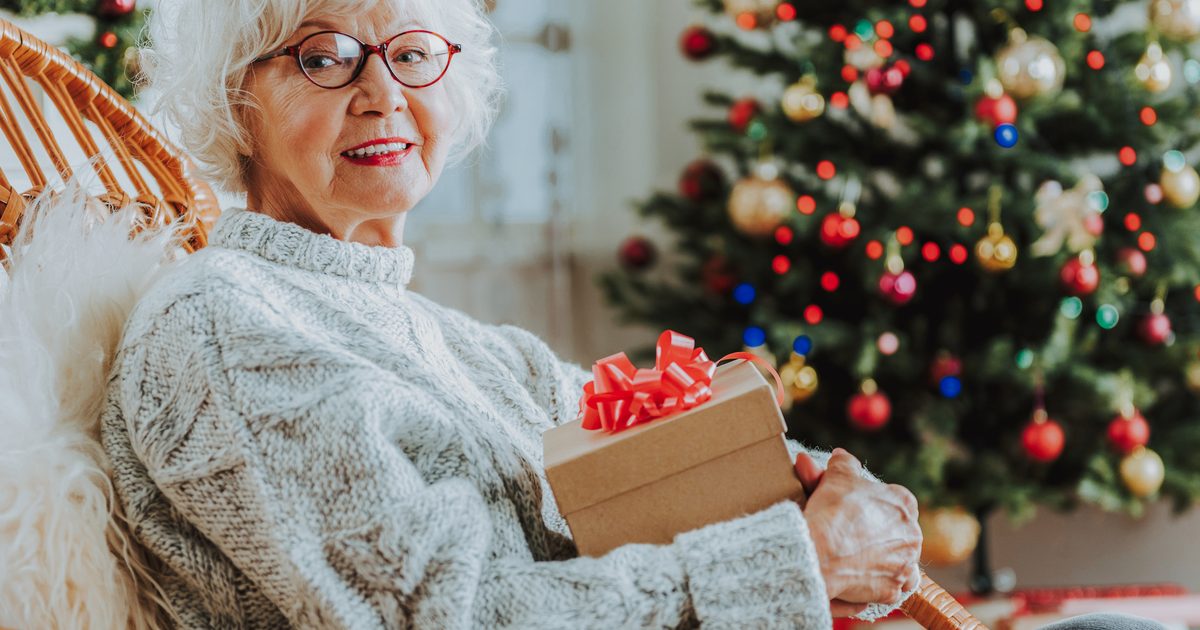 Una donna anziana sorridente con un pacchetto regalo davanti all'albero di Natale